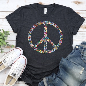 May include: A dark heather grey t-shirt with a peace sign made of colorful leaves. The peace sign is centered on the shirt. The shirt is laid out on a white wooden surface with a pair of white sneakers and blue jeans.