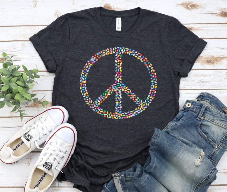 May include: A dark heather grey t-shirt with a peace sign made of colorful leaves. The peace sign is centered on the shirt. The shirt is laid out on a white wooden surface with a pair of white sneakers and blue jeans.