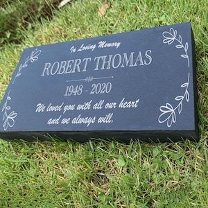 May include: A rectangular, dark gray memorial plaque with white engraved text and floral accents. The inscription reads "In Loving Memory ROBERT THOMAS 1948-2020 We loved you with all our heart and we always will."