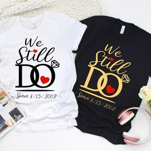 Peut inclure: Deux t-shirts blancs et noirs avec le texte "We Still Do" et la date "Since 8/15/2002". Les t-shirts présentent un design de bague stylisé avec un diamant et un cœur.