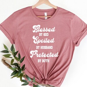 K&ouml;nnte beinhalten: Ein mauvefarbenes T-Shirt mit dem Text "Blessed by God, Spoiled by Husband, Protected by Both" in Wei&szlig;.
