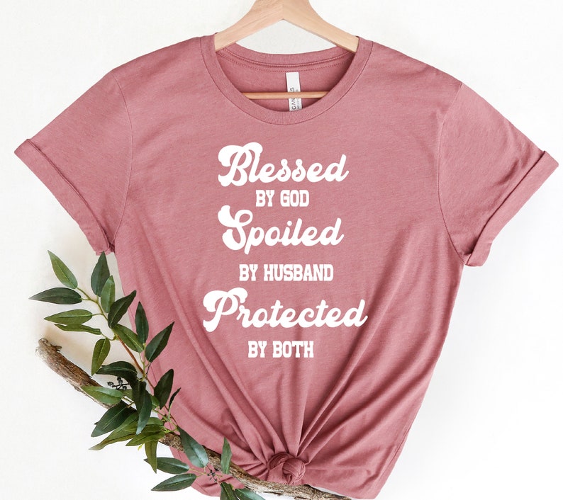 K&ouml;nnte beinhalten: Ein mauvefarbenes T-Shirt mit dem Text "Blessed by God, Spoiled by Husband, Protected by Both" in Wei&szlig;.