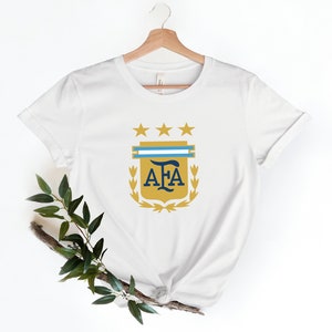May include: White t-shirt with a gold crest featuring three stars, a blue and white stripe, and the letters "AFA" in blue.