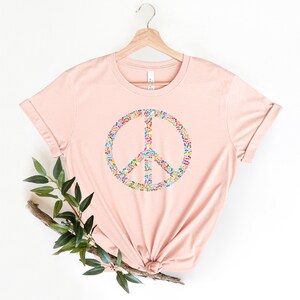 May include: A pink t-shirt with a peace sign made of colorful leaves. The peace sign is centered on the shirt.