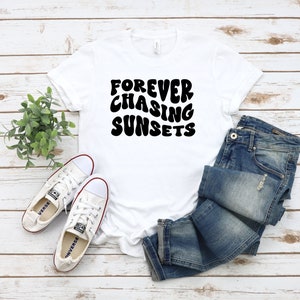 May include: White t-shirt with black text that reads "FOREVER CHASING SUNSETS" in a retro font. The shirt is laid flat on a woodgrain surface with a pair of white sneakers and blue jeans.