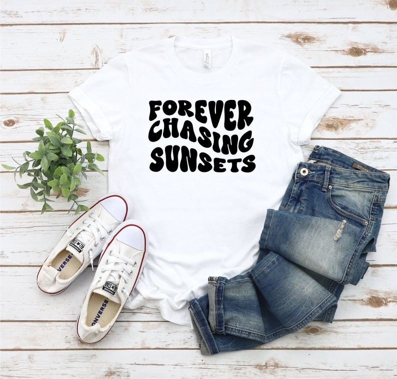 May include: White t-shirt with black text that reads "FOREVER CHASING SUNSETS" in a retro font. The shirt is laid flat on a woodgrain surface with a pair of white sneakers and blue jeans.