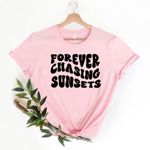 May include: Pink t-shirt with the text "FOREVER CHASING SUNSETS" in black, retro-style lettering.