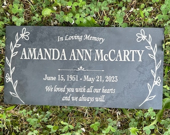 May include: A black slate memorial plaque with white lettering. The plaque reads "In Loving Memory Amanda Ann McCarty June 15, 1951 - May 21, 2023 We loved you with all our hearts and we always will."