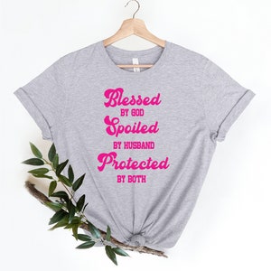 K&ouml;nnte beinhalten: Graues T-Shirt mit dem Text "Blessed by God, Spoiled by Husband, Protected by Both" in rosa.