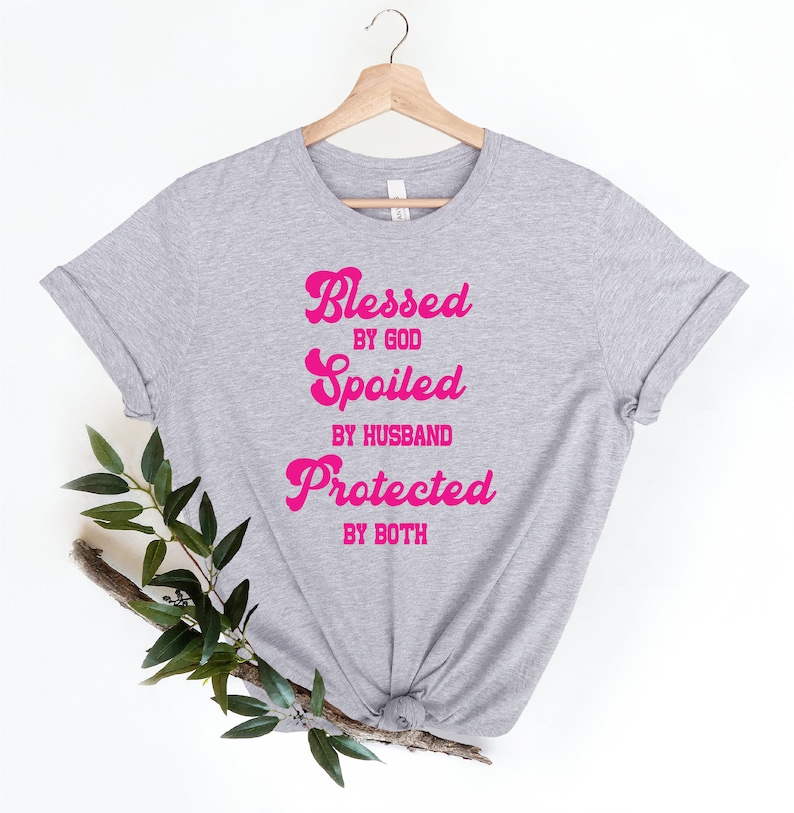 K&ouml;nnte beinhalten: Graues T-Shirt mit dem Text "Blessed by God, Spoiled by Husband, Protected by Both" in rosa.