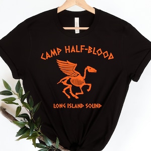 Può includere: Maglietta nera con uno scheletro arancione di un cavallo alato e il testo "Camp Half-Blood Long Island Sound".