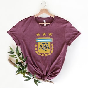 May include: A maroon t-shirt with a gold crest featuring three stars, a blue and white stripe, and the letters "AFA" in navy blue.