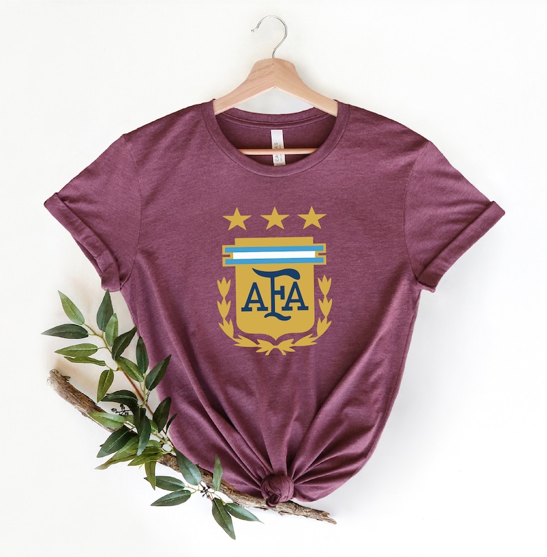 May include: A maroon t-shirt with a gold crest featuring three stars, a blue and white stripe, and the letters "AFA" in navy blue.