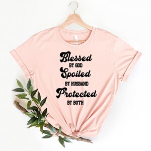 K&ouml;nnte beinhalten: Ein rosa T-Shirt mit dem Text "Blessed by God, Spoiled by Husband, Protected by Both" in Schwarz.