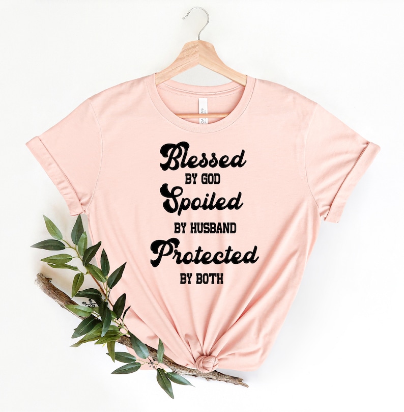K&ouml;nnte beinhalten: Ein rosa T-Shirt mit dem Text "Blessed by God, Spoiled by Husband, Protected by Both" in Schwarz.
