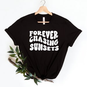 May include: Black t-shirt with white text that reads "FOREVER CHASING SUNSETS" in a retro font. The shirt is hanging on a wooden hanger against a white background.