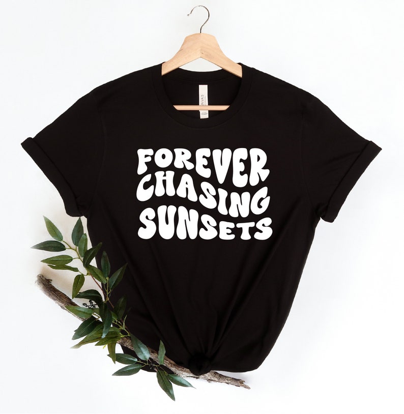 May include: Black t-shirt with white text that reads "FOREVER CHASING SUNSETS" in a retro font. The shirt is hanging on a wooden hanger against a white background.