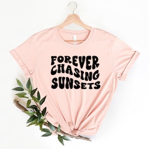 May include: A pink t-shirt with the text "FOREVER CHASING SUNSETS" in black, retro-style lettering.
