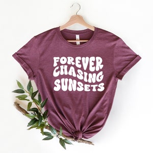 May include: A maroon t-shirt with the text "FOREVER CHASING SUNSETS" in white, stylized lettering. The shirt is hanging on a wooden hanger against a white background.