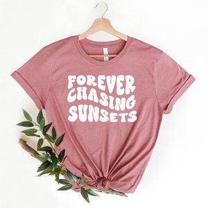 May include: A mauve colored t-shirt with the text "FOREVER CHASING SUNSETS" in white, stylized lettering. The shirt is hanging on a wooden hanger.