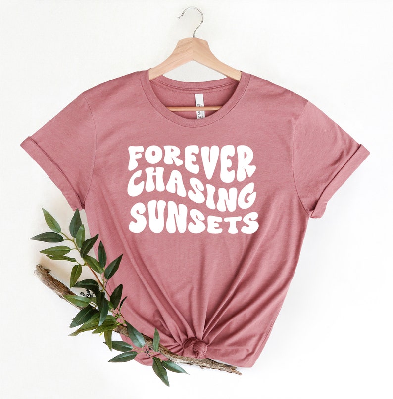 May include: A mauve colored t-shirt with the text "FOREVER CHASING SUNSETS" in white, stylized lettering. The shirt is hanging on a wooden hanger.