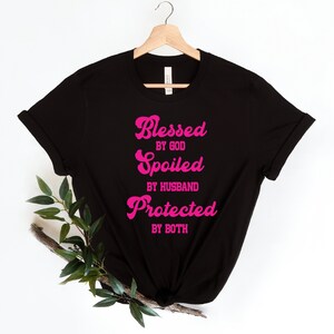 K&ouml;nnte beinhalten: Schwarzes T-Shirt mit pinkfarbenem Text, der "Gesegnet von Gott, Verw&ouml;hnt vom Ehemann, Gesch&uuml;tzt von beiden" lautet.