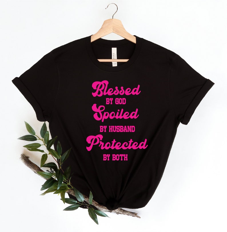 K&ouml;nnte beinhalten: Schwarzes T-Shirt mit pinkfarbenem Text, der "Gesegnet von Gott, Verw&ouml;hnt vom Ehemann, Gesch&uuml;tzt von beiden" lautet.