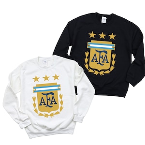 May include: Two long-sleeved sweatshirts with the Argentinian Football Association (AFA) logo. The sweatshirt on the left is white with a gold logo. The sweatshirt on the right is black with a gold logo.