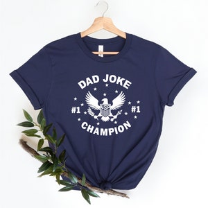 Op de afbeelding: Donkerblauw T-shirt met een witte afbeelding van een adelaar met de woorden "Dad Joke Champion" en "#1" aan weerszijden van de adelaar.