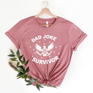 Op de afbeelding: Een stoffig roze T-shirt met een witte afbeelding waarop "Dad Joke Survivor" staat met een adelaar en sterren.