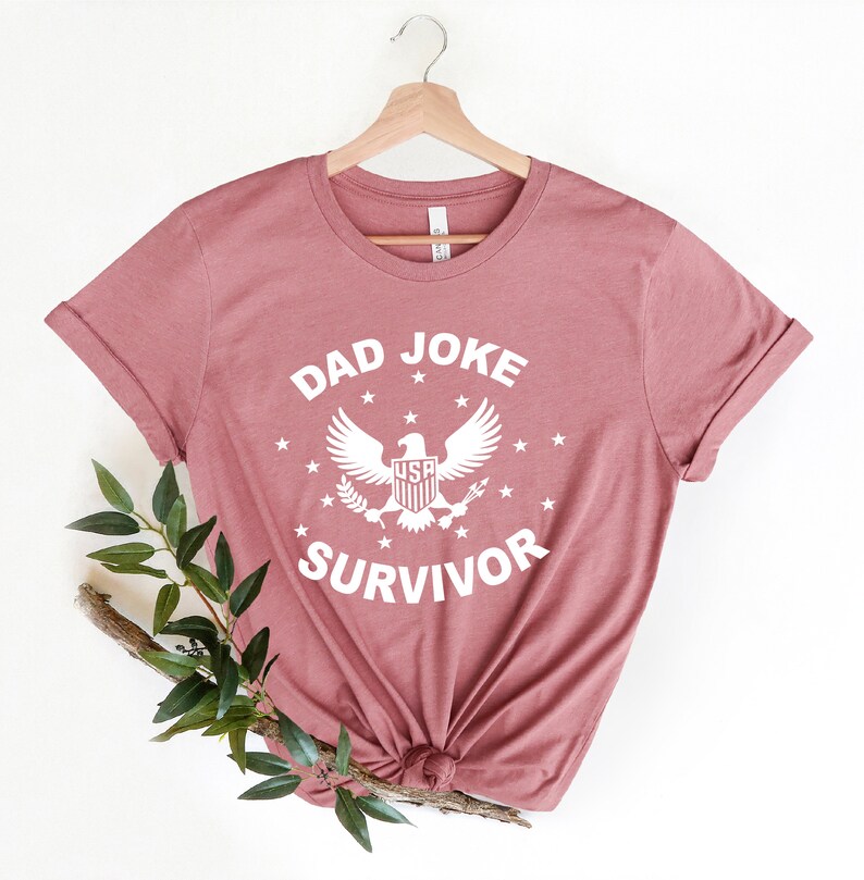 Op de afbeelding: Een stoffig roze T-shirt met een witte afbeelding waarop "Dad Joke Survivor" staat met een adelaar en sterren.