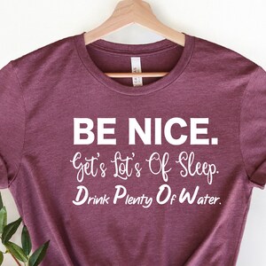 Peut inclure: Un t-shirt marron avec un texte blanc qui dit "BE NICE. Get's Lot's Of Sleep. Drink Plenty Of Water."