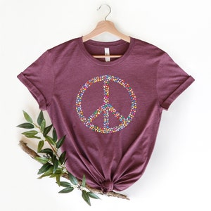 May include: A maroon t-shirt with a peace sign made of colorful leaves. The peace sign is centered on the shirt.