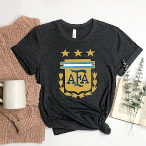 May include: A dark gray t-shirt with a gold and blue crest featuring the letters "AFA" and three stars. The crest is surrounded by a laurel wreath.
