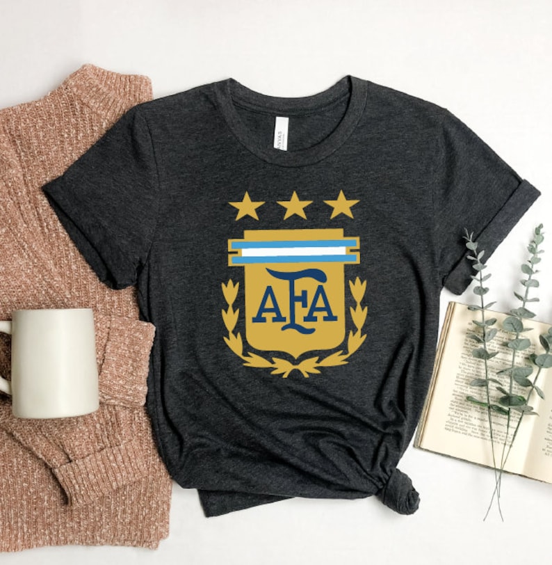 May include: A dark gray t-shirt with a gold and blue crest featuring the letters "AFA" and three stars. The crest is surrounded by a laurel wreath.