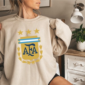 May include: A beige sweatshirt with a gold and blue Argentinian Football Association (AFA) logo. The logo features a shield with three stars, a blue and white stripe, and the letters "AFA" in a gold font.