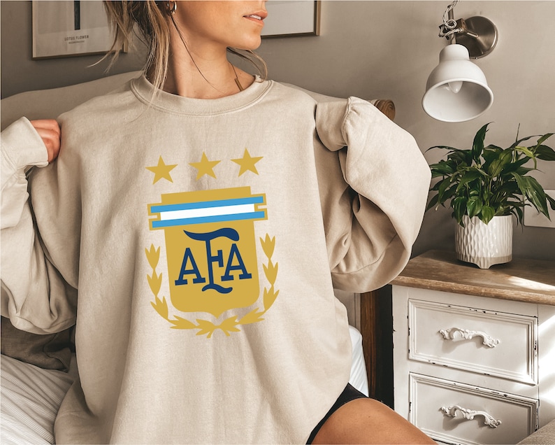 May include: A beige sweatshirt with a gold and blue Argentinian Football Association (AFA) logo. The logo features a shield with three stars, a blue and white stripe, and the letters "AFA" in a gold font.