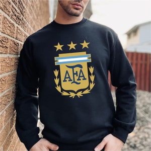 May include: Black sweatshirt with a gold and blue Argentinian soccer team crest. The crest features three gold stars, a blue and white stripe, and the letters "AFA" in gold.