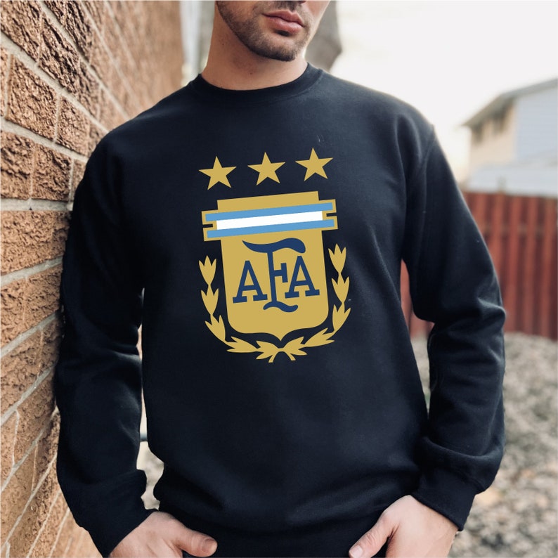 May include: Black sweatshirt with a gold and blue Argentinian soccer team crest. The crest features three gold stars, a blue and white stripe, and the letters "AFA" in gold.