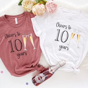 Könnte beinhalten: Zwei weiße T-Shirts mit dem Text "Cheers to 10 years" und einer Grafik von Champagnergläsern. Die Shirts liegen auf einer weißen, flauschigen Oberfläche mit einer Flasche Champagner.