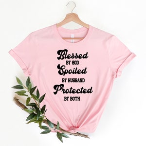 K&ouml;nnte beinhalten: Rosafarbenes T-Shirt mit schwarzem Text, der "Gesegnet von Gott, Verw&ouml;hnt vom Ehemann, Gesch&uuml;tzt von beiden" lautet.
