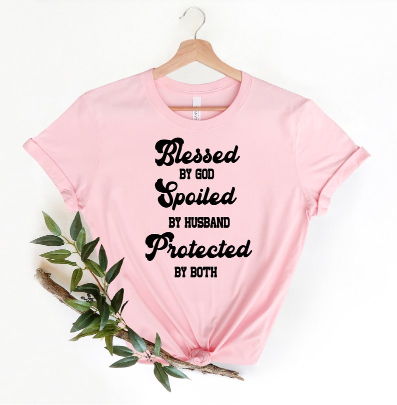 K&ouml;nnte beinhalten: Rosafarbenes T-Shirt mit schwarzem Text, der "Gesegnet von Gott, Verw&ouml;hnt vom Ehemann, Gesch&uuml;tzt von beiden" lautet.