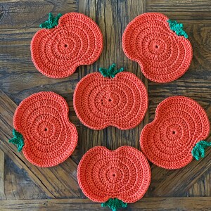 May include: Six crocheted pumpkin-shaped coasters in a bright orange color with green stems. The coasters are arranged in a circle on a wooden surface.