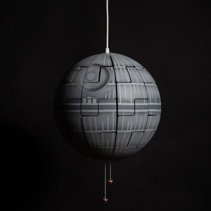 Op de afbeelding: Een grijze, bolvormige hanglamp die lijkt op de Death Star uit Star Wars. De lamp heeft een segmentontwerp met gedetailleerde panelen en een wit snoer. Twee kleine koperkleurige decoratieve elementen hangen eronder.