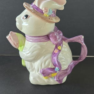 Puede incluir: Una tetera de cerámica blanca con forma de conejo, adornada con un sombrero y una bufanda. El sombrero es marrón con una banda morada y detalles florales. La bufanda y el asa son moradas con detalles florales. El pico tiene forma de flor.