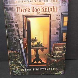 Peut inclure: Couverture du livre "Three Dog Knight" de Johnnie Alexander. Elle présente un mur en pierre avec une porte ouverte révélant une table avec un bonsaï et des figurines de chiens. Le titre est en grandes lettres rouges.