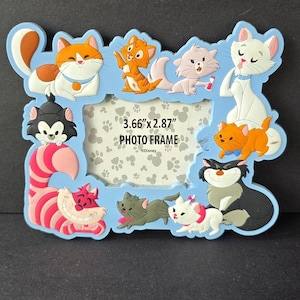 Può includere: Una cornice blu chiaro con personaggi di gatti dei cartoni animati. La cornice ha un'apertura rettangolare con il testo "3.66" x 2.87" PHOTO FRAME". La cornice è decorata con varie illustrazioni di gatti in arancione, bianco, grigio e nero.