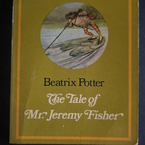 Op de afbeelding: Een vintage boek met de titel "The Tale of Mr. Jeremy Fisher" van Beatrix Potter. De olijfgroene kaft toont een illustratie van een vissende kikker. De titel en de naam van de auteur staan in sierlijke letters.