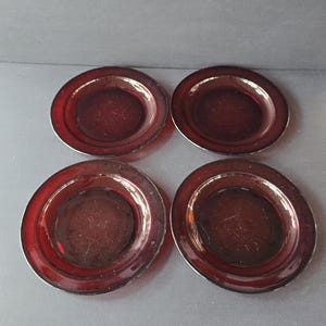 Op de afbeelding: Vier kleine, ronde, robijnrode glazen borden met een licht verhoogde rand. De borden hebben een eenvoudig, elegant ontwerp en zijn gerangschikt op een donkergrijze ondergrond. De borden zijn waarschijnlijk voor het serveren van kleine porties.
