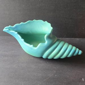 May include: A matte turquoise ceramic bowl shaped like a conch shell. The vessel features a wide, scalloped opening that tapers into a spiraled, ribbed tail. The surface has a smooth, uniform finish with subtle color gradients throughout the piece.